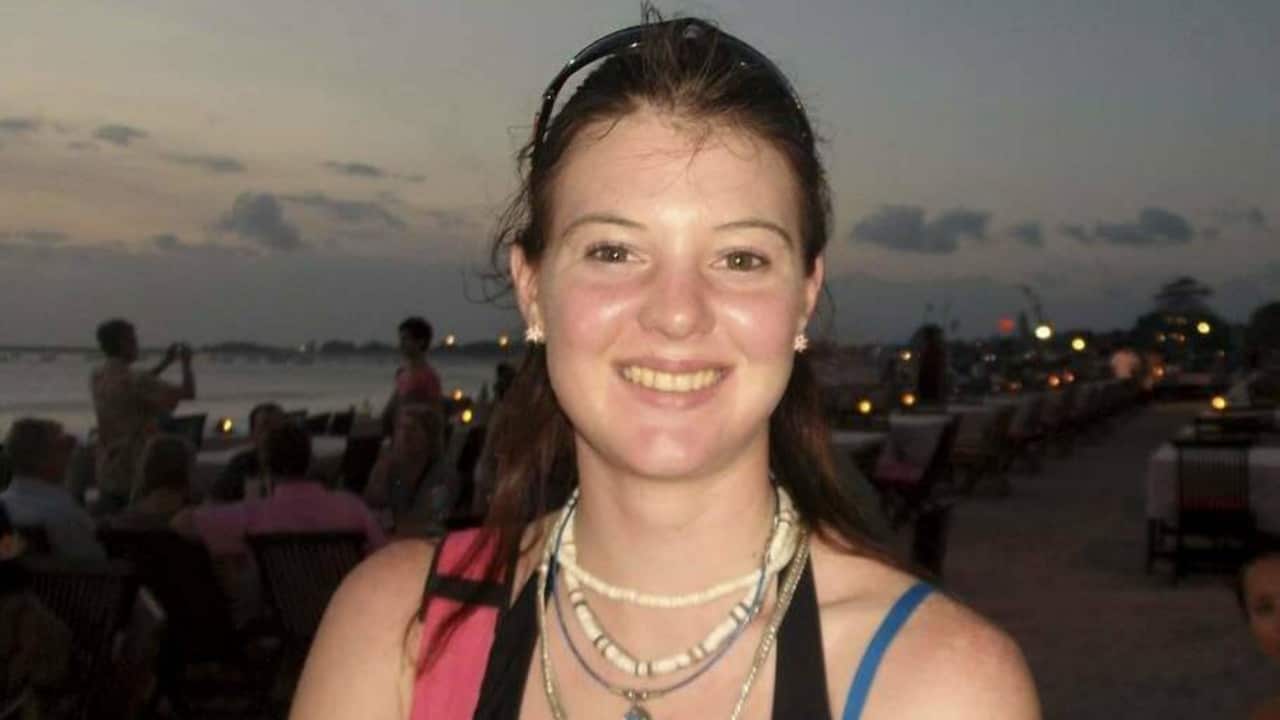 a young woman with a brown ponytail and shell necklaces smiles at the camera on a crowded beach at night time