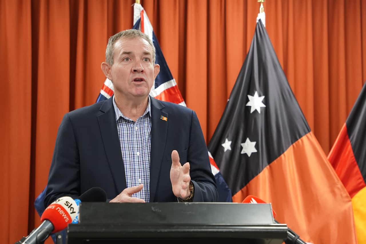 A man in suit speaking at a podium indoors. There is an Australian and a Northern Territory flag behind him