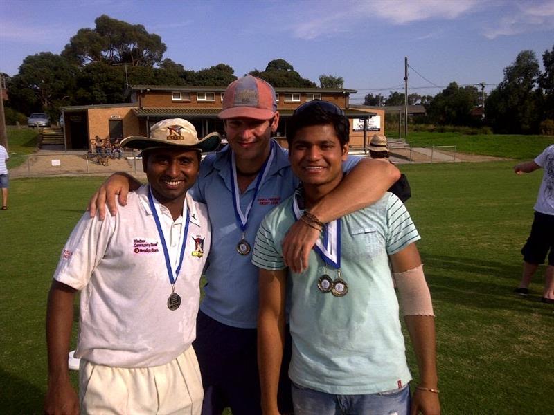 Three men wear medals and smile for a photo on a green cricket oval with a clubhouse in the background.