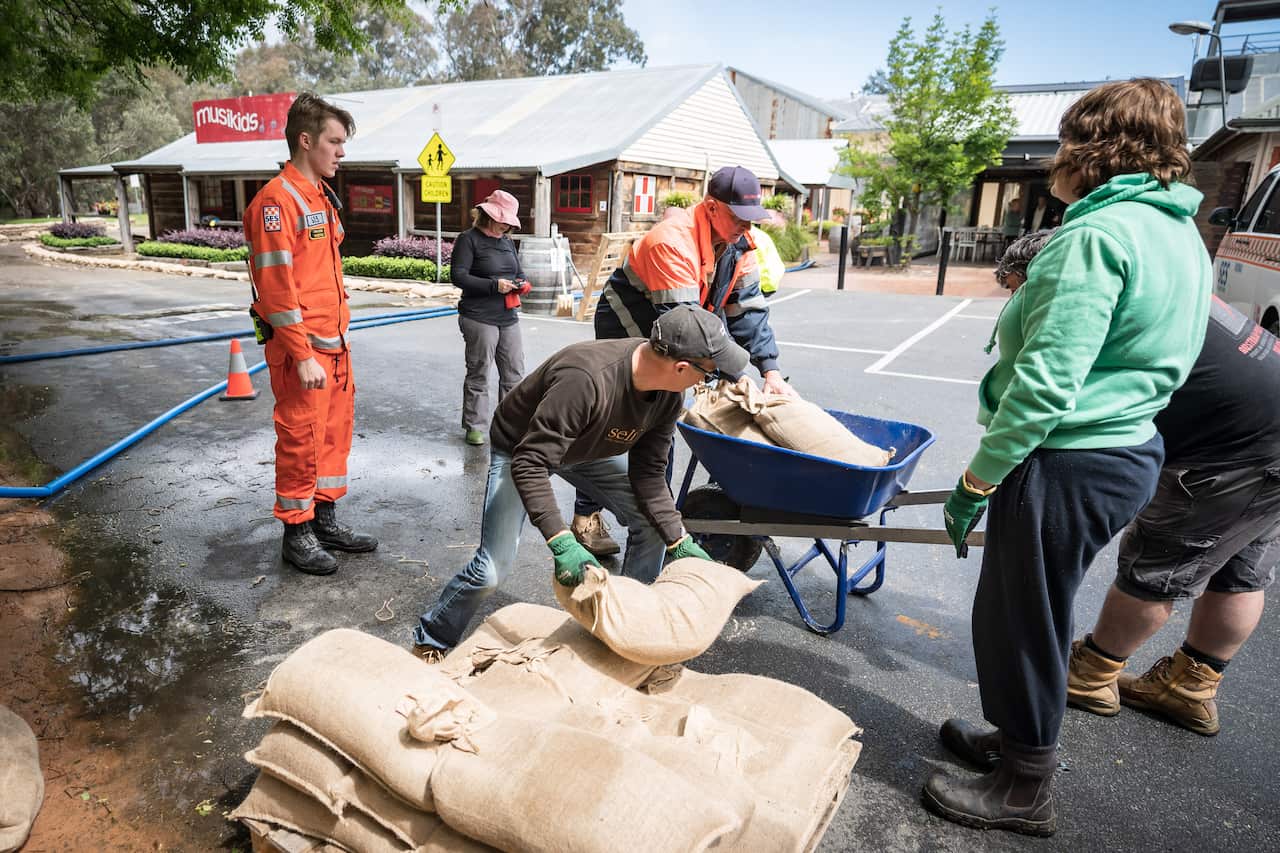 A man standing in yellow overalls near a man adding a sandbag to a pile of sandbags and another man removing a sandbag from a wheelbarrow on a street. There is a woman standing just behind them