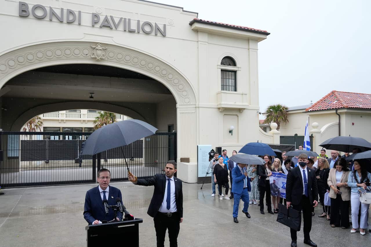 A man in a navy suit speaking before a lectern outside the Bondi Pavilion. To his left, people crowd under umbrellas as they watch on. 