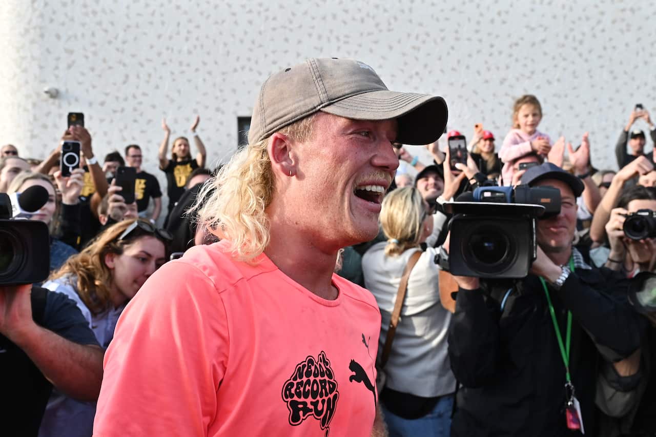 Un hombre con un salmonete rubio y una camisa rosa sonríe mientras está rodeado por una multitud de personas y fotógrafos.