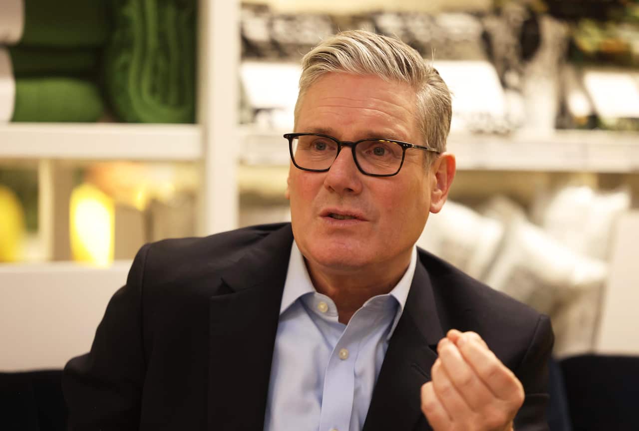 Keir Starmer, wearing a black jacket and blue shirt, waving his hand slightly.