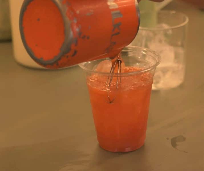 An orange drink is poured from a jug into a glass filled with ice, sitting on a table.
