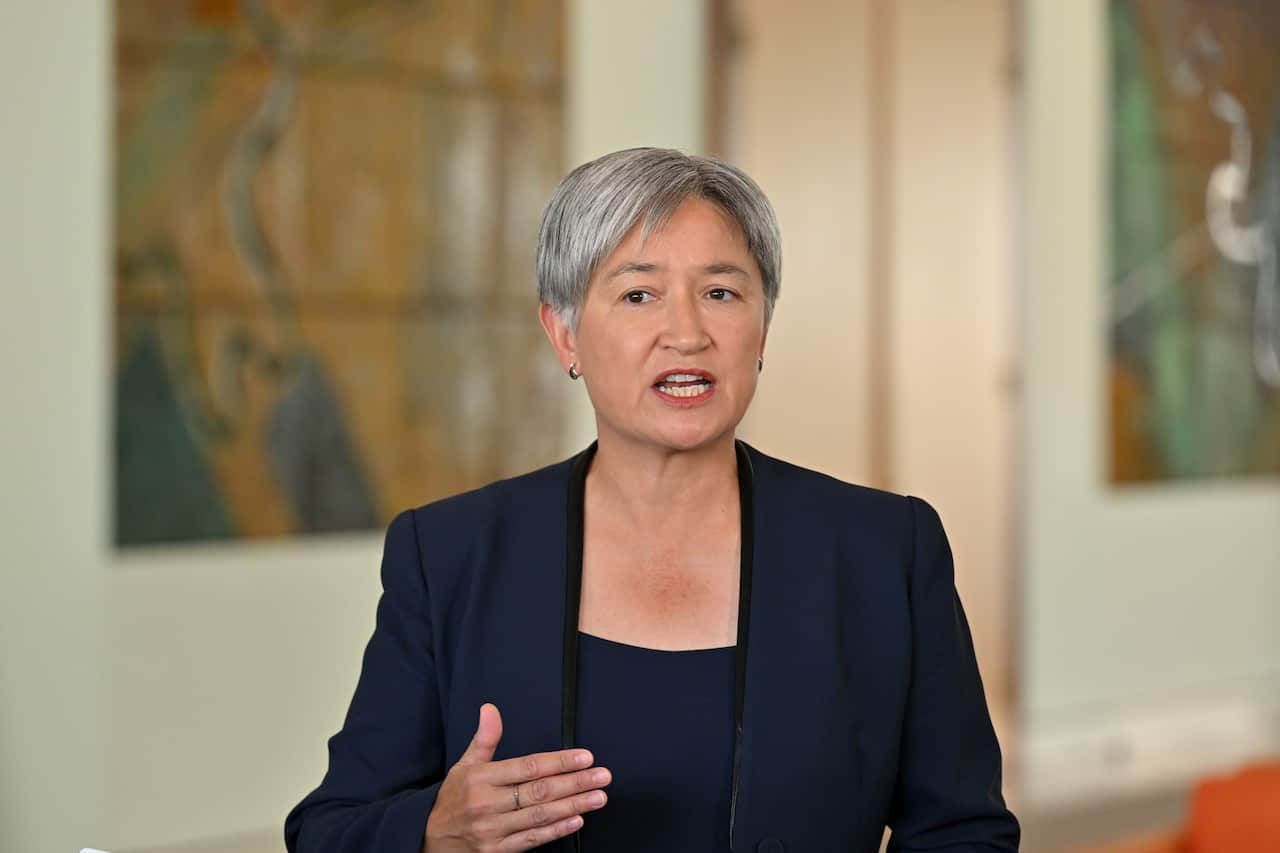 Australian Foreign Minister Penny Wong, with short grey hair and wearing a navy blue blazer, gestures with her hand while speaking in front of a blurred background.