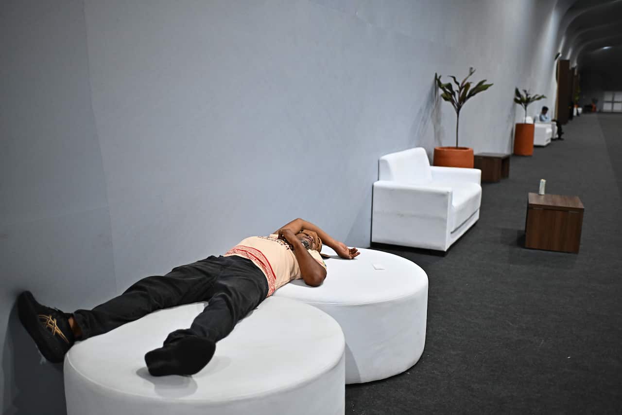 A man in an orange shirt sleeps on two white chairs in a grey room.