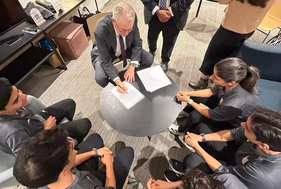 A man signing documents sitting at a table, while five women sit around him.