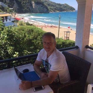 A man in a white T-shirt is sitting near a beach.