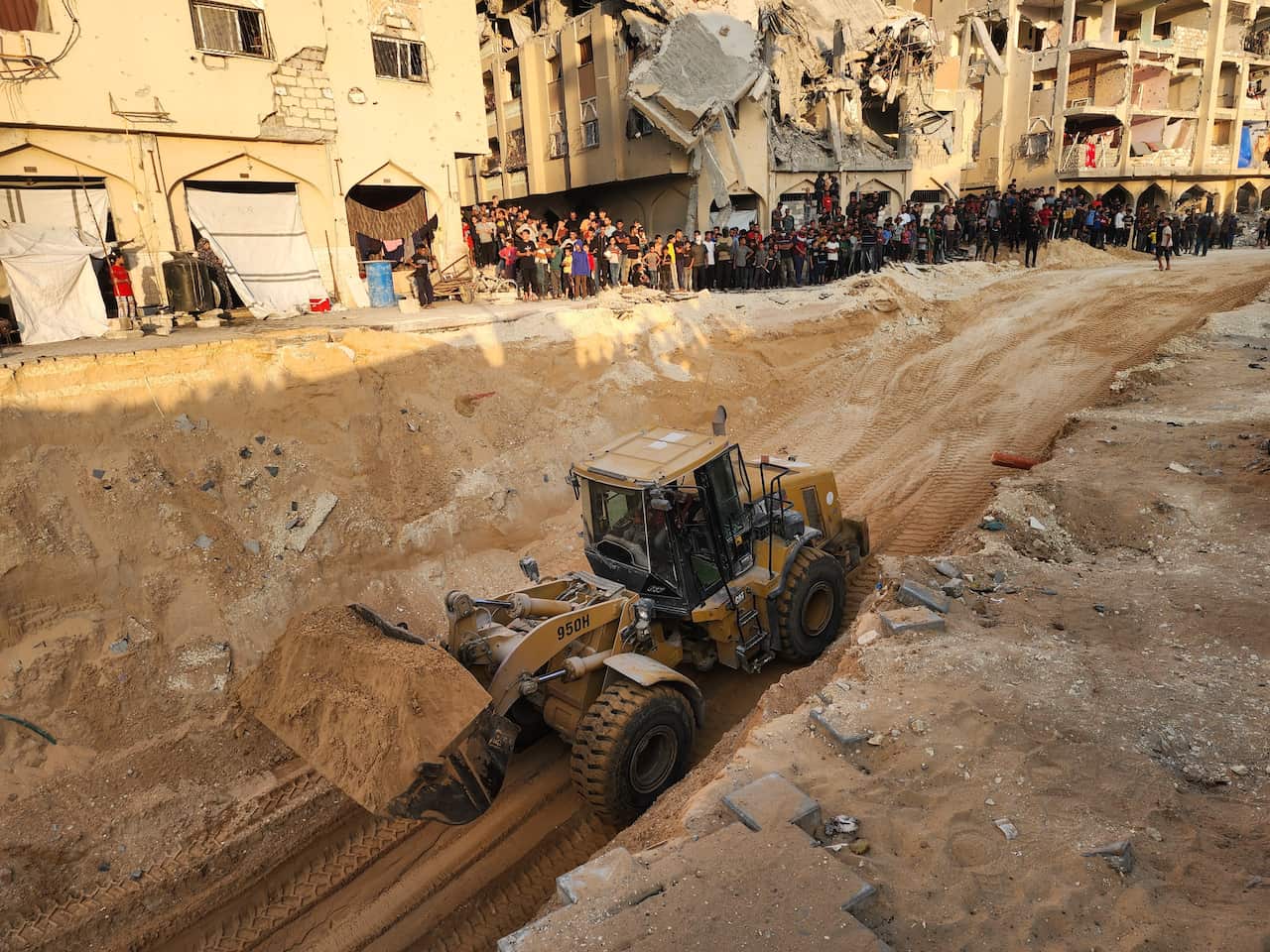 An excavator digs up dirt among rubble and as a crowd of Palestinians watch on