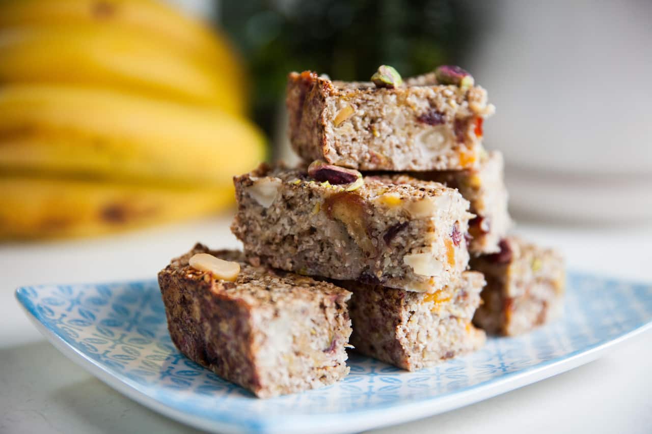 Rectangular pieces of a cake-like bake, studded with fruit and nuts, sit on a patterned blue plate. Out of focus bananas can be seen in the background. 