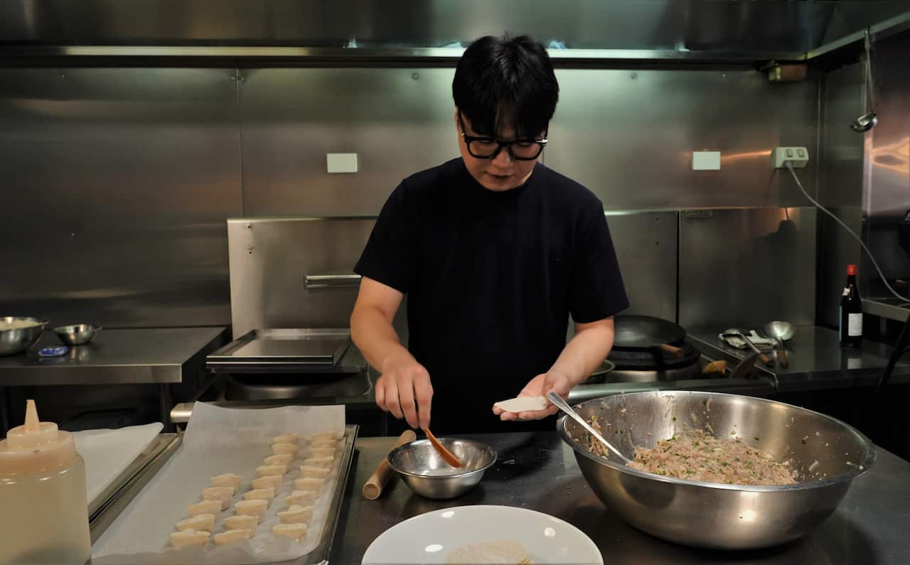 A Korean man in a black t-shirt, making dumplings. On his left is a large metal bowl half full of a meat-based dumpling filling, the man is holding a dumpling wrapper preparing to place filling in it, and to his left is a baking tray with three rows of dumplings.