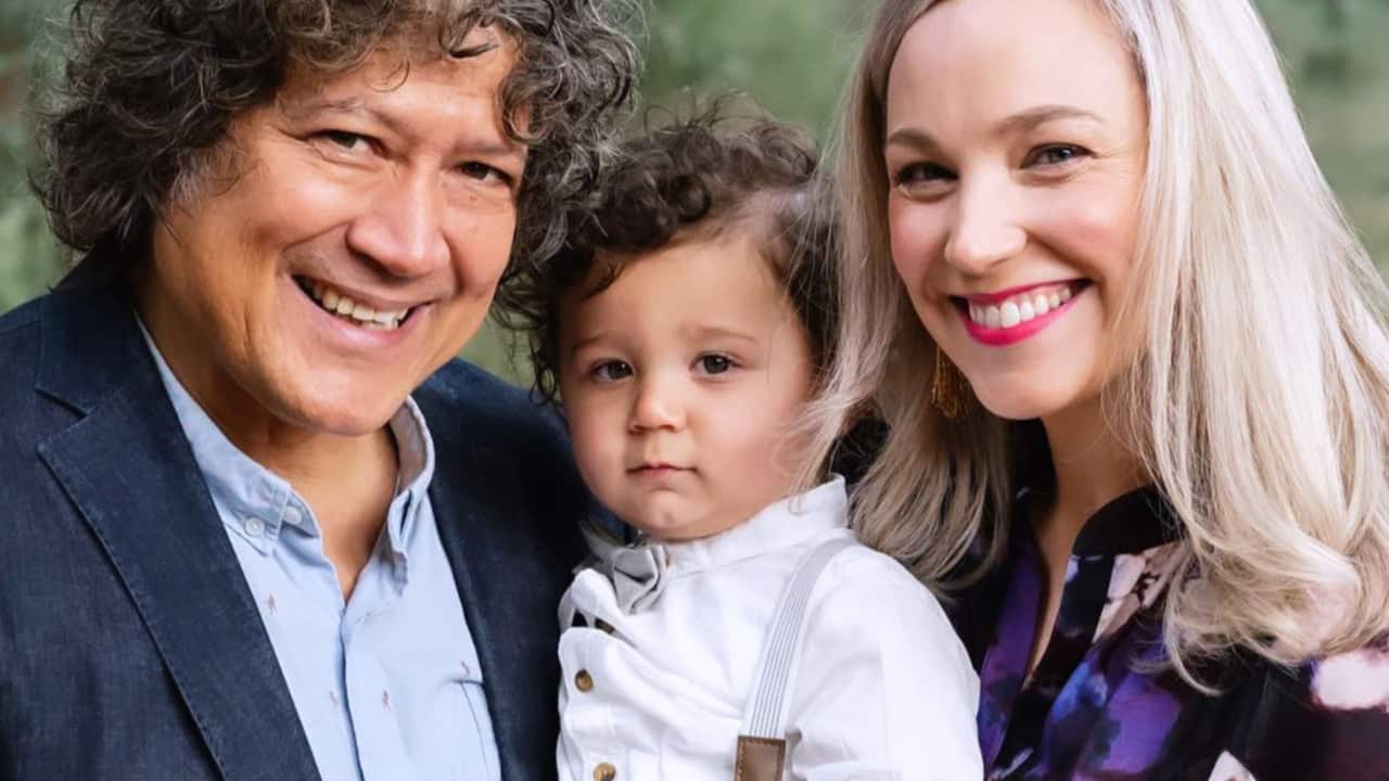 a family portrait of a man with dark curly hair in a suit and a woman with long blonde hair and their toddler son who has suspenders and a white collared shirt and brown curly hair