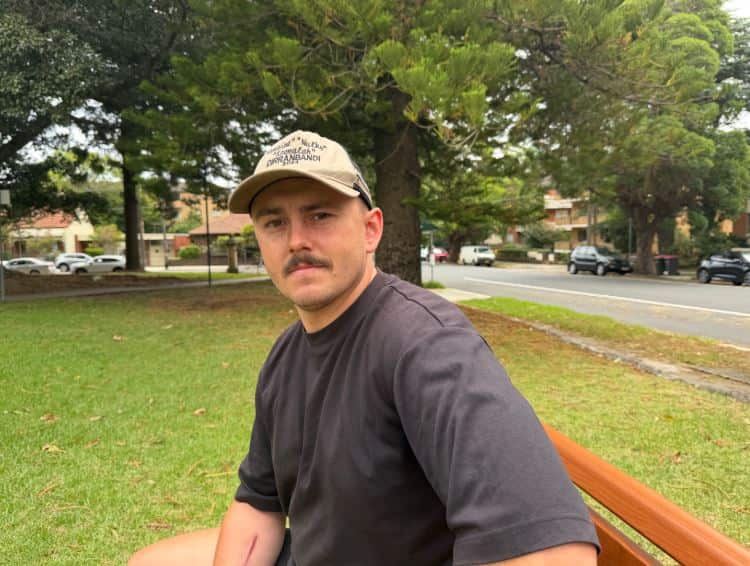 A man in a black t shirt and cap and sitting on a park bench