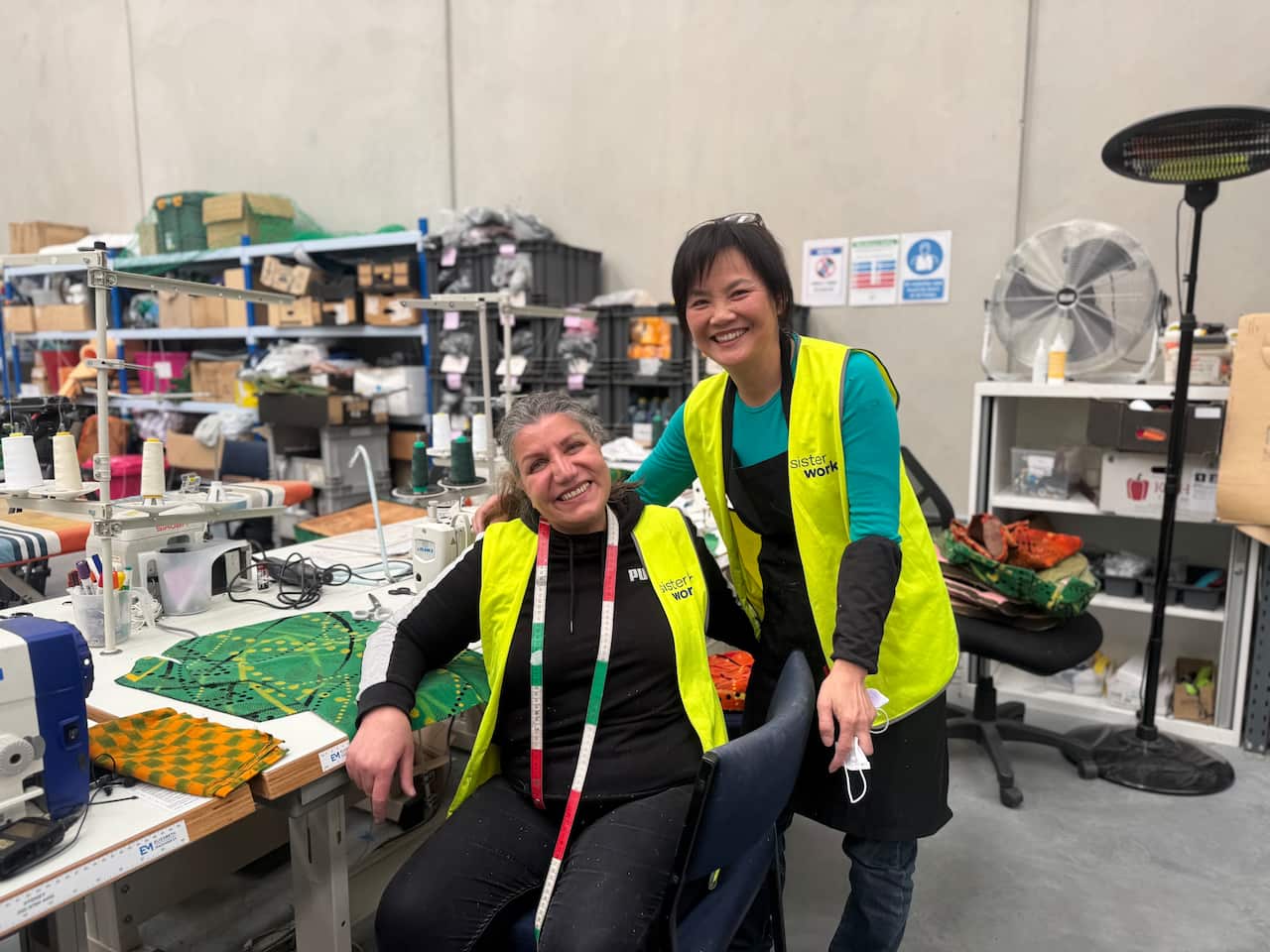 A woman is standing next to another woman sitting on a chair. They are inside a sewing workshop, wearing yellow hi-vis vests.