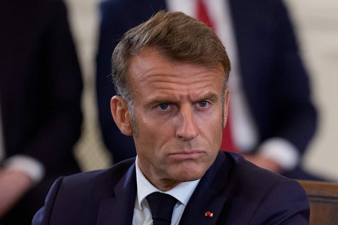 Emmanuel Macron sitting in a chair. The bodies of two people in suits can be seen behind him.