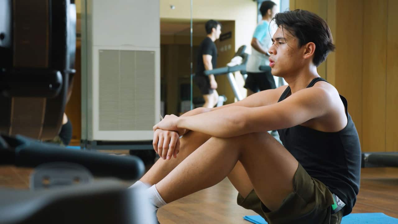 A young man in dark workout clothes sitting on the gym floor and catching his breath during a workout break