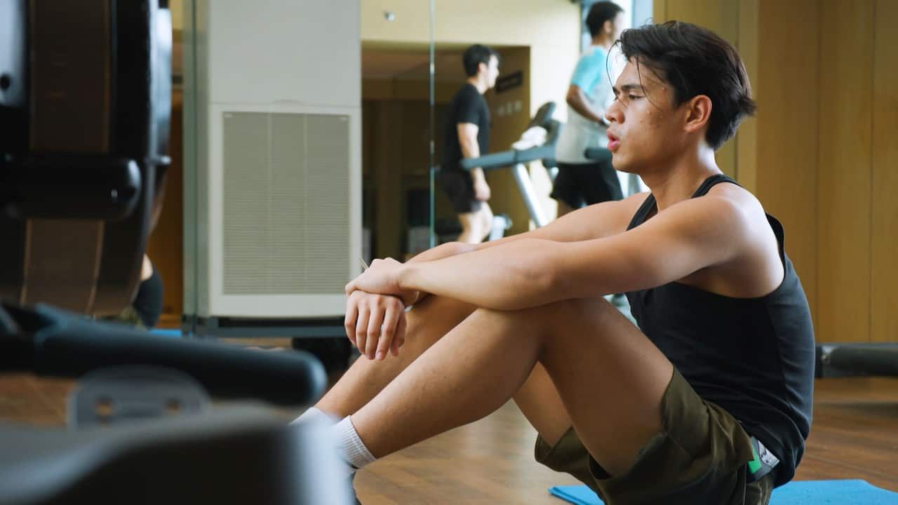 A young man in dark workout clothes sitting on the gym floor and catching his breath during a workout break 