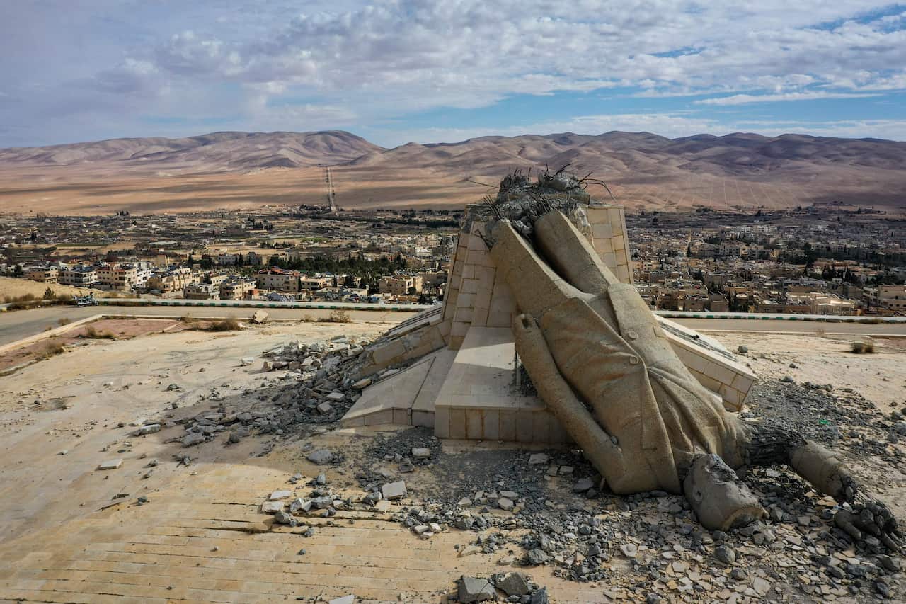 A toppled stone statue lying in rubble overlooking a desert town and distant hills.