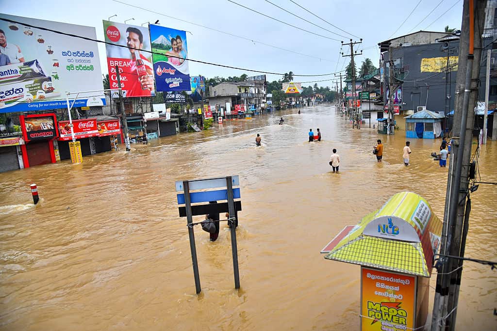 People in a wide street lined with buildings. The street is flooded with brown water