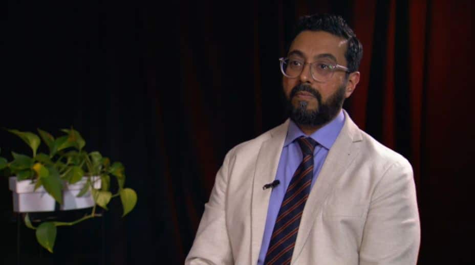 A man in an off-white suit jacket, sitting inside.