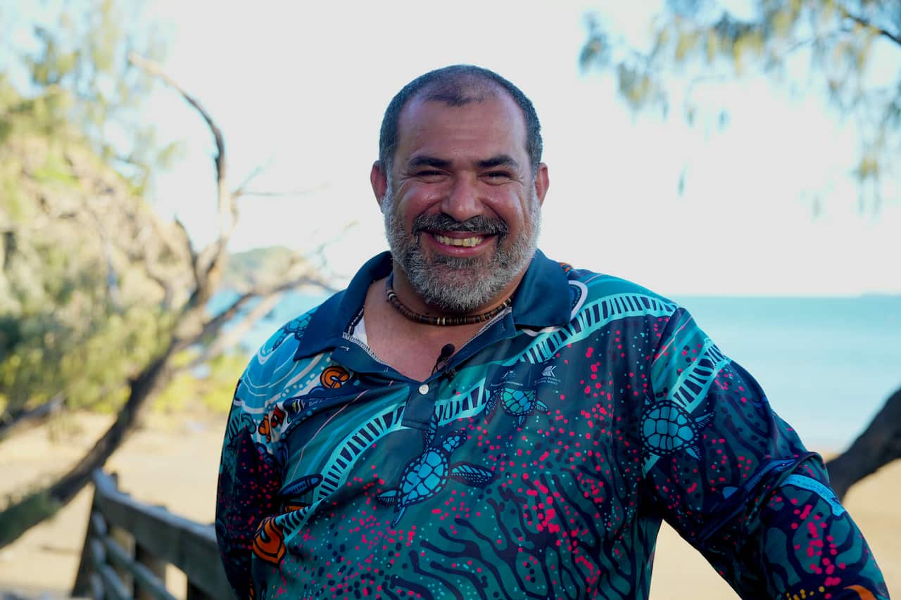 Man smiling wearing blue long sleeve shirt with beach behind him