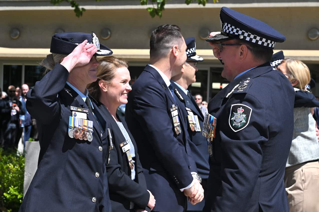 New Australian Federal Police Commissioner Krissy Barrett farewells her predecessor with a salute. Both are dressed in police uniform at a ceremony with other police officials in the background.