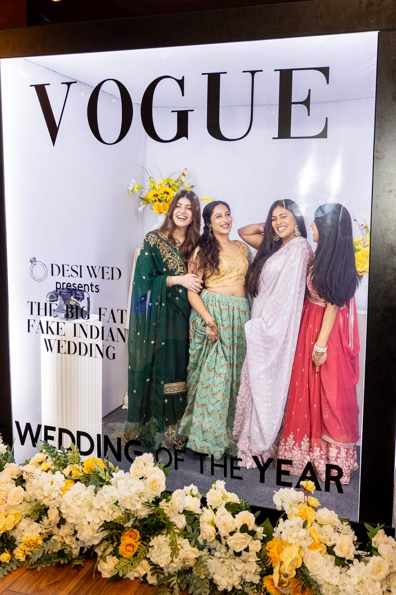 Quatro mulheres vestidas com trajes de casamento indianos posam juntas dentro de uma grande moldura de vidro projetada para parecer a capa de uma revista Vogue.
