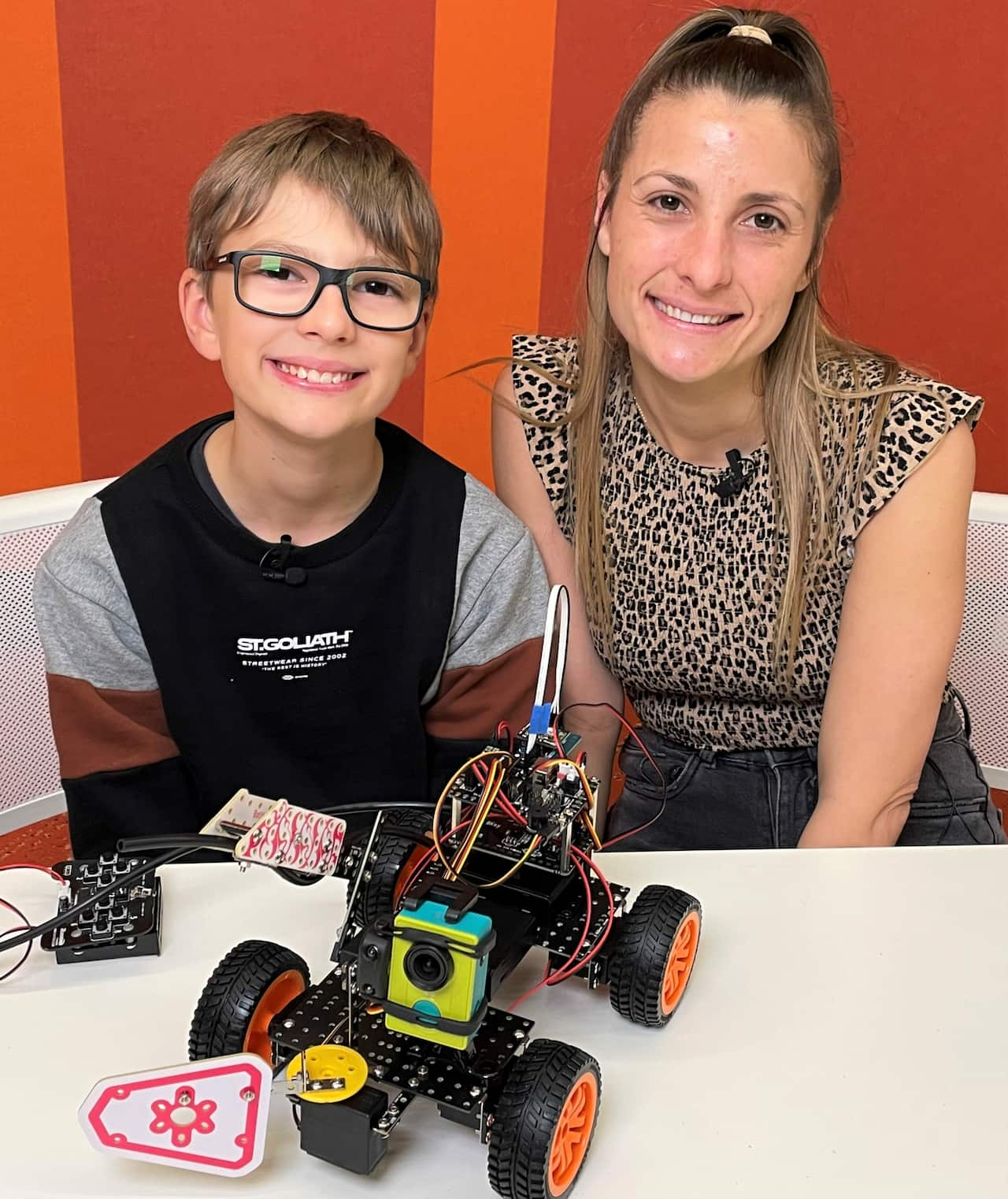 A boy sitting with his mother. A robot with four wheels is on a table in front of them