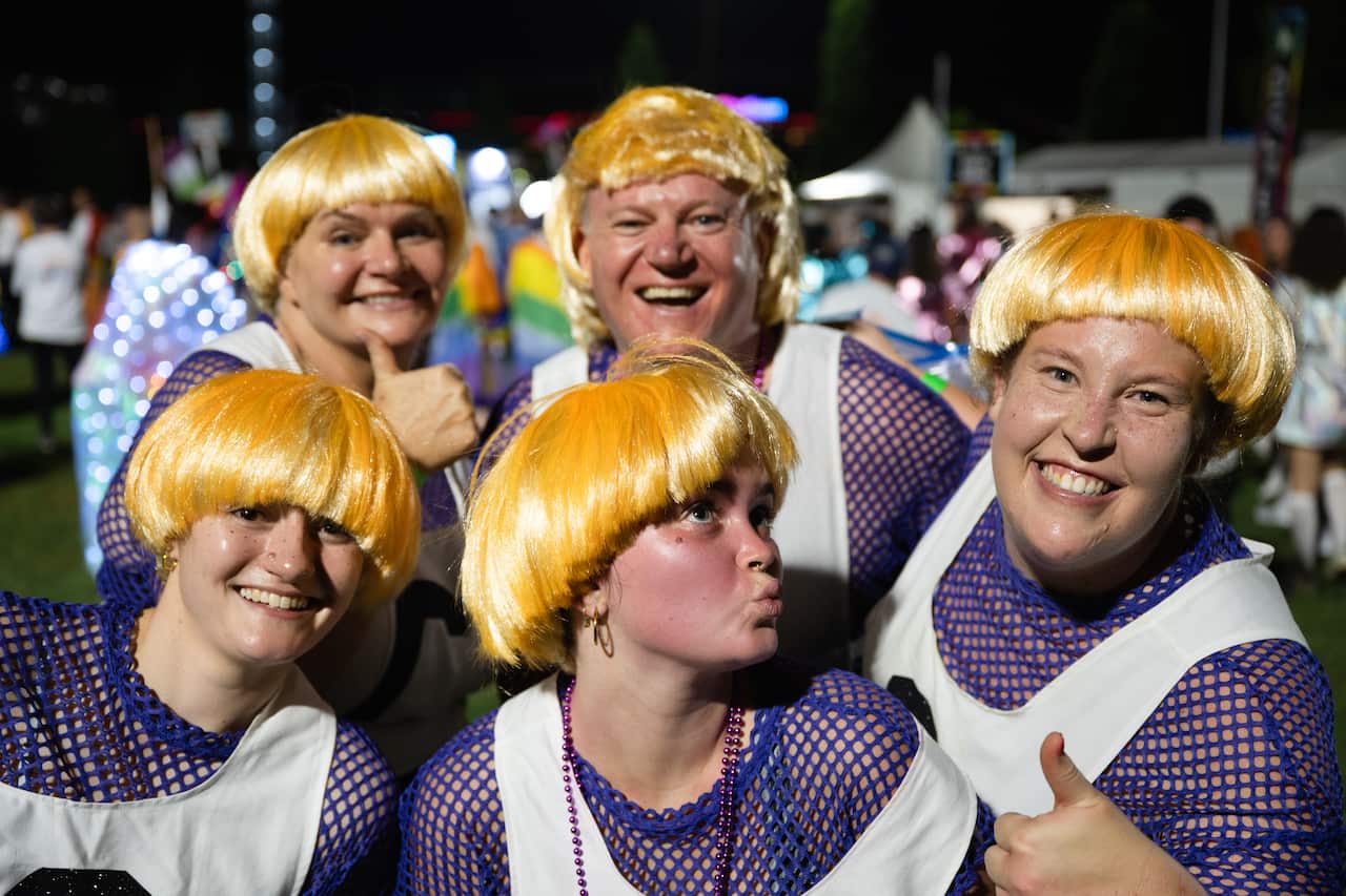 Un grupo de personas sonrientes con pelucas rubias y camisetas de netball levantan el pulgar hacia la cámara.