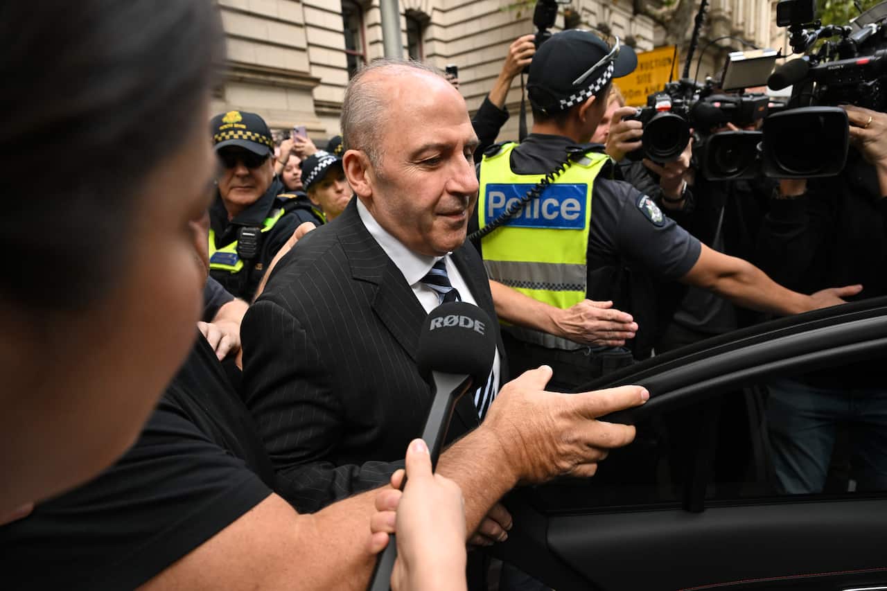 A man in a black suit is surrounded by a crowd, including police and members of the media.