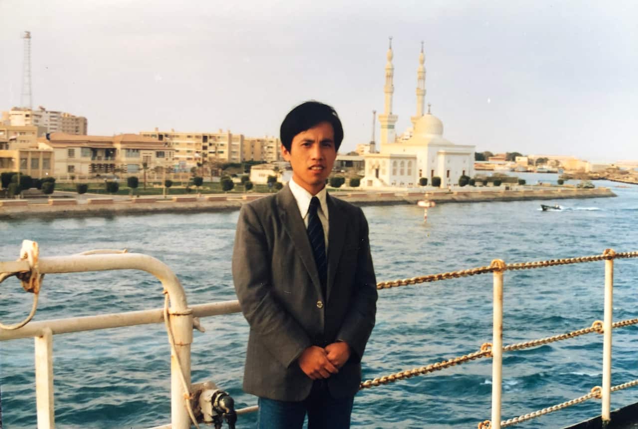 A man on board a ship, with water behind him. 