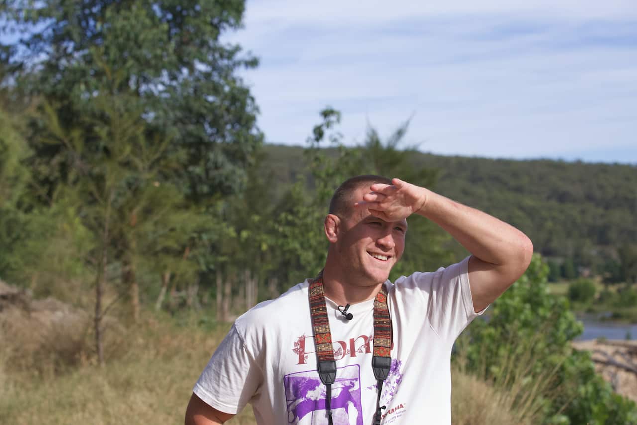 A man in a white t-shirt stands in nature, blocking his eyes from the sun.