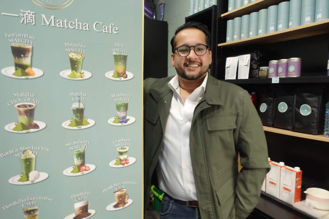 A man posing beside a menu of matcha drinks printed on a wall. He is wearing a green jacket and white shirt