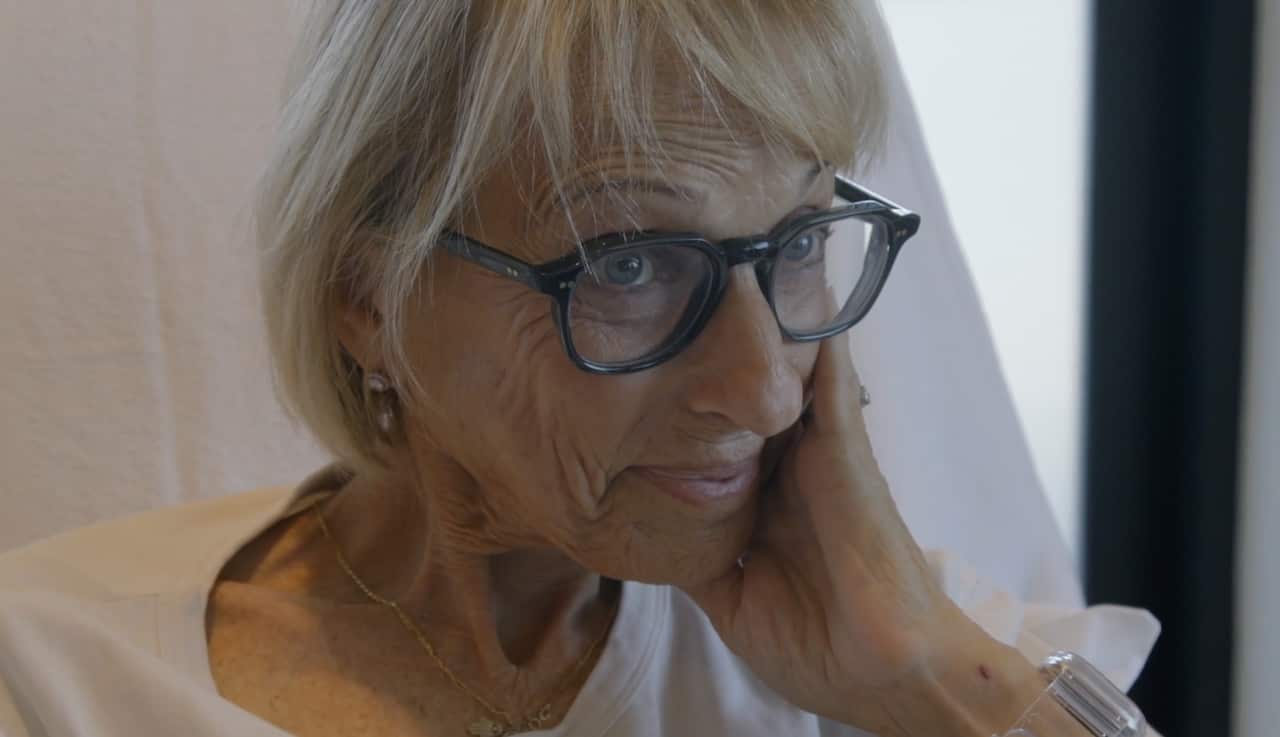 A close-up of an older woman sitting in a chair and smiling. She has her left hand against her cheek.