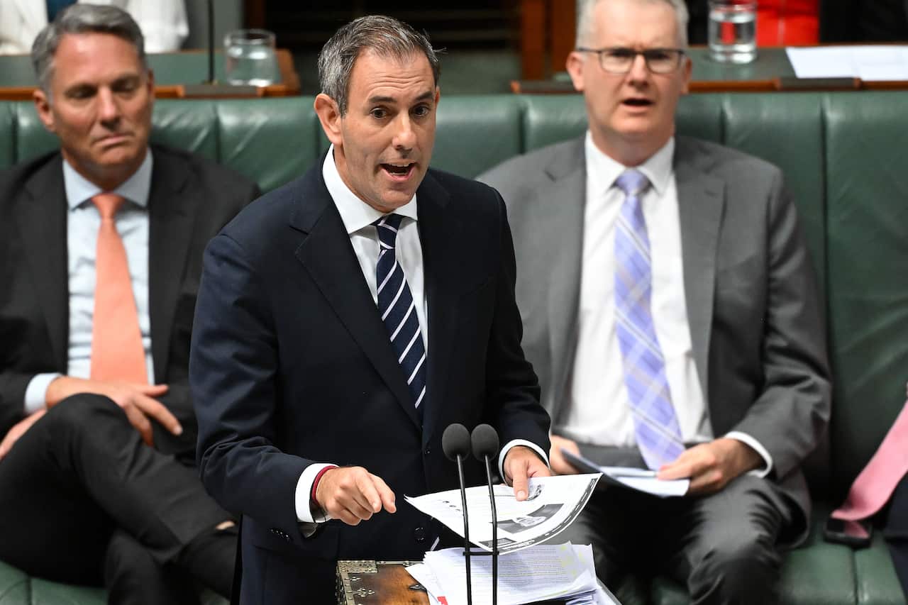 A man in a black suit and striped tie speaks while standing in front of a microphone with other MPs sitting behind him. 