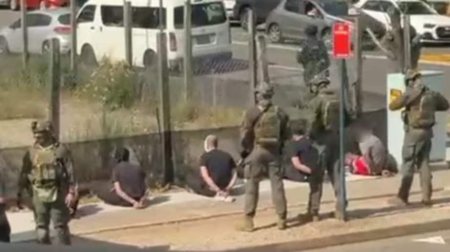 Heavily armed police in camoflauge uniforms stand beside men in who are seated on a footpath with their zip-tied behind their backs