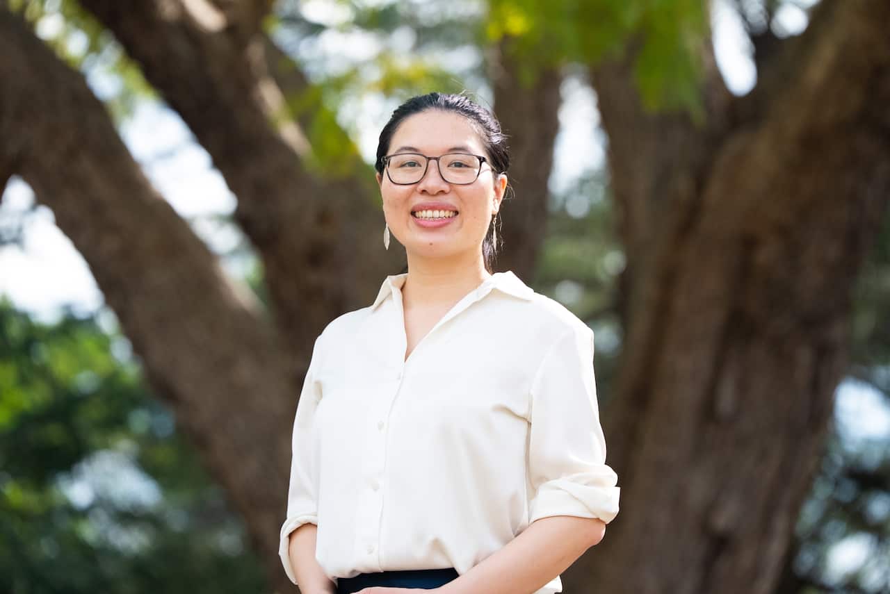 A woman wearing a white shirt is smiling as she stands in front of a tree.