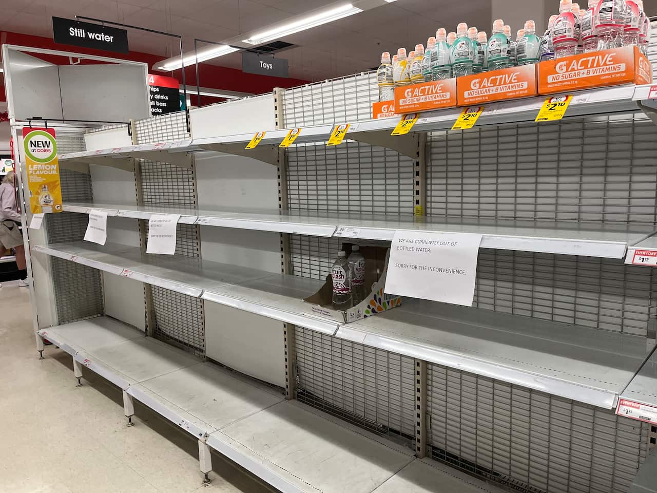 Empty supermarket shelves in the bottled water aisle with only a few drinks left and signs saying the store is out of water.