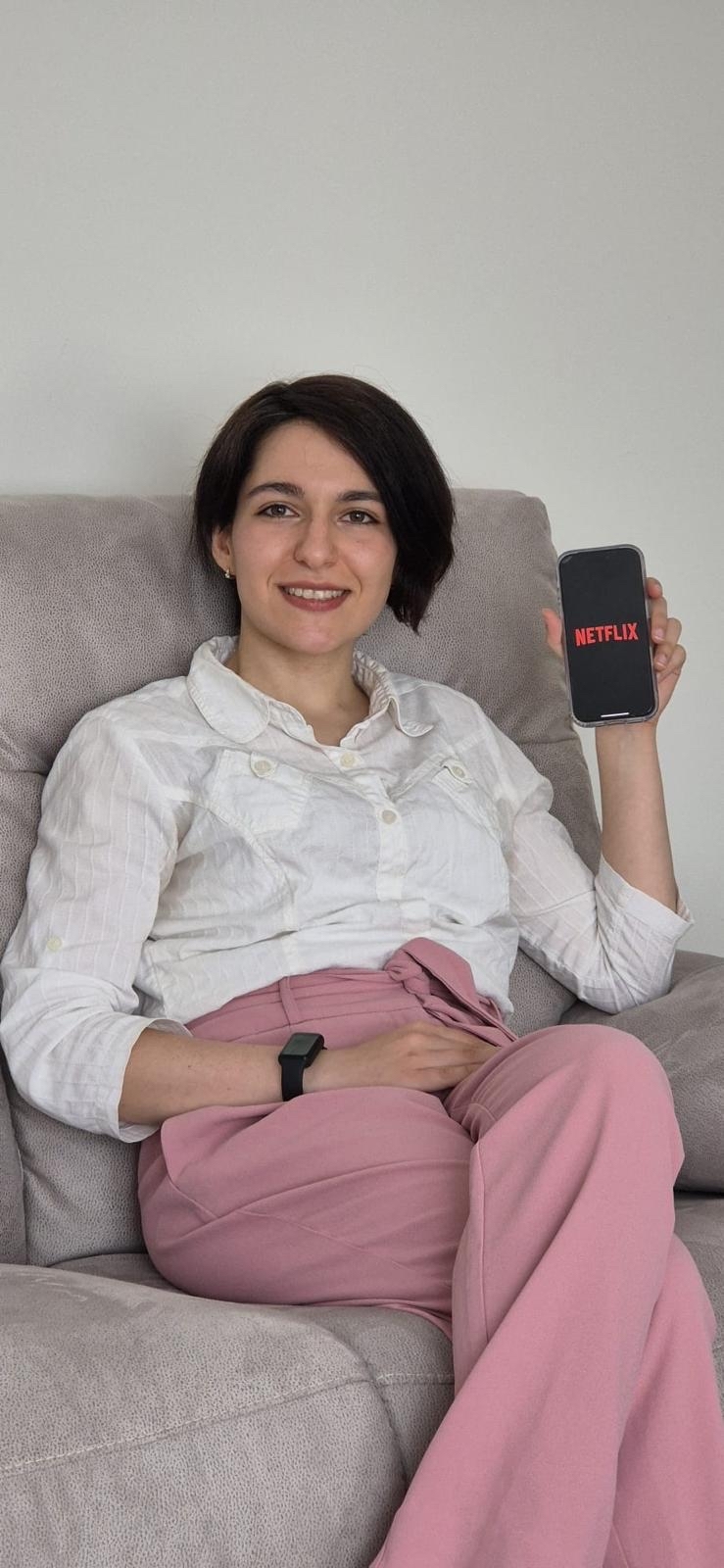 A woman sitting on a sofa holds up a phone, which shows the Netflix logo