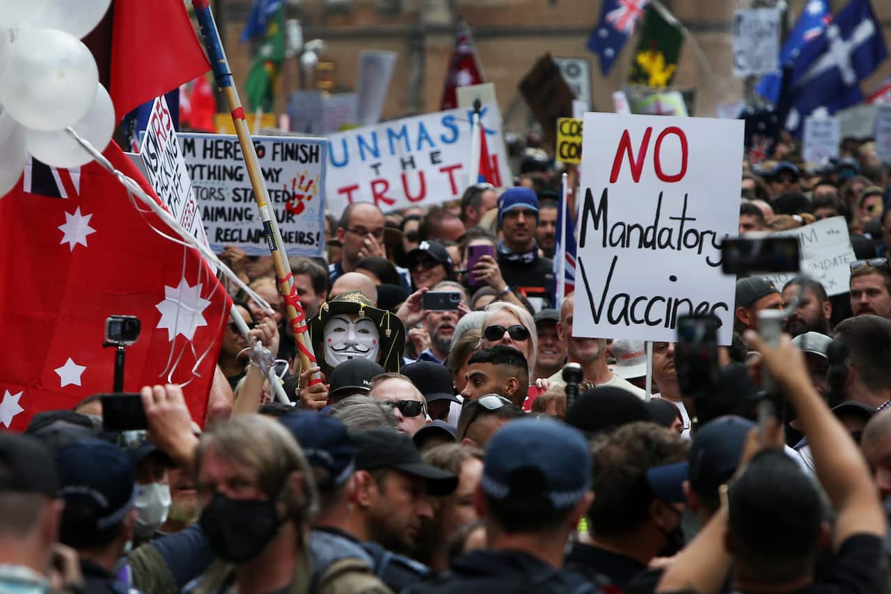 A crowd of protesters, some holding signs, including one that reads "NO mandatory vaccines" and another that says "Unmask the truth."