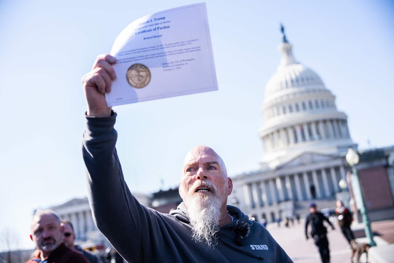 Un hombre de mediana edad, calvo y con barba blanca, sostiene un certificado de indulto presidencial frente al Capitolio de Estados Unidos.