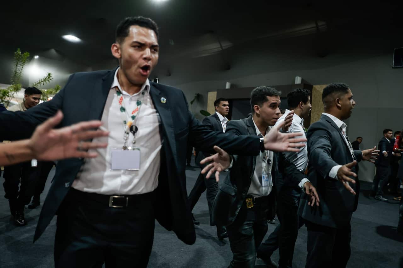 A row of men in suits and lanyards hold their arms out as they move through a large, dark indoor space.