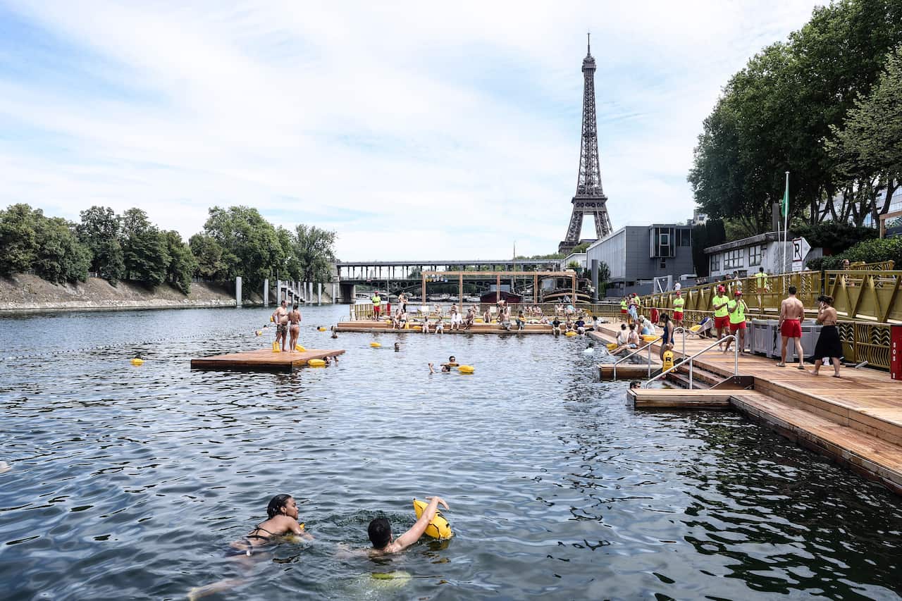 Swimmers in the river Seine use banana-shaped floating devices, while some stand or sit on pontoons and lifeguards in brightly coloured outfits can be seen to the side. In the background is the Eiffel Tower. 