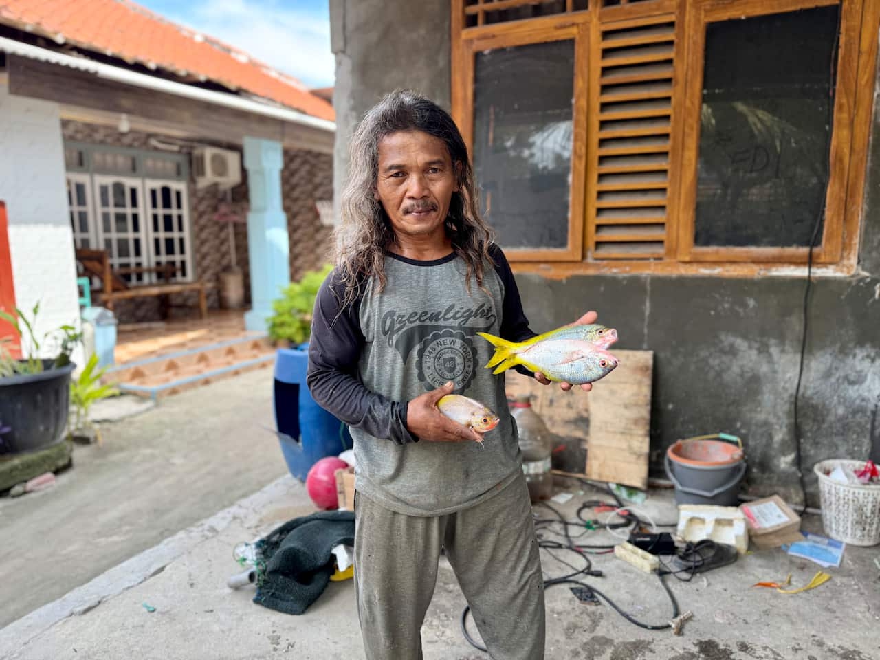 Um homem vestindo calça cinza claro e uma camiseta cinza de mangas compridas com mangas pretas segura dois pequenos peixes prateados com cauda amarela na mão esquerda. Ele também segura um pequeno peixe na mão direita.