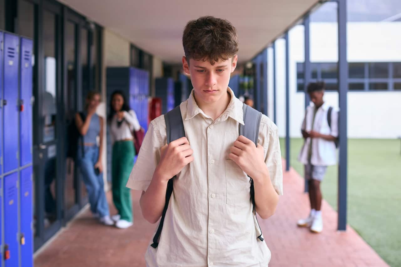 A sad or worried-looking teenage boy with a backpack walks down a school hallway past a row of blue lockers, with other students visible but out of focus in the background.
