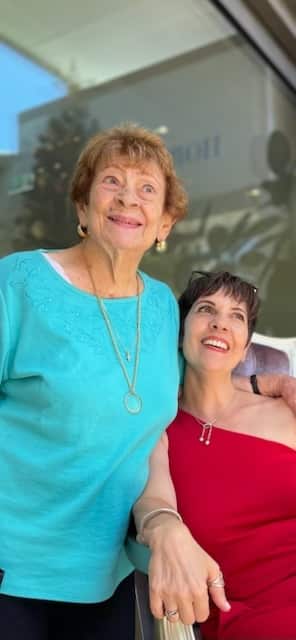 A woman in a red dress sitting next to an older woman in an aqua top, who is standing. Both are smiling