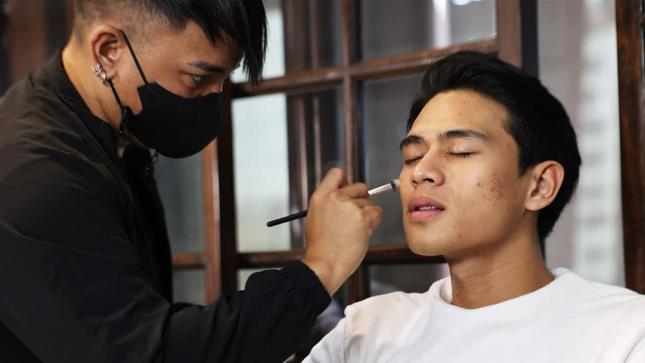 A makeup artist wearing a black face mask applying makeup with a brush on a young man's face 