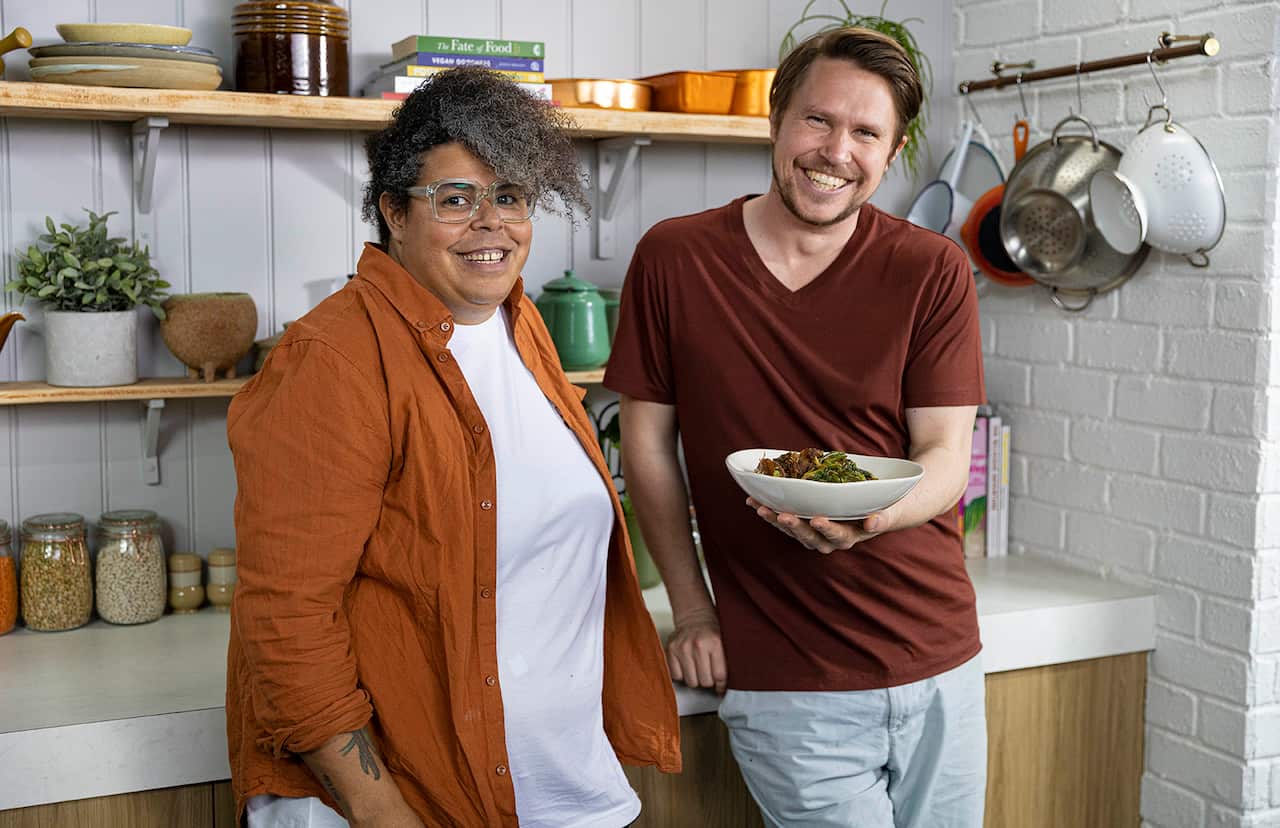 A man and a woman stand in a kitchen. He's holding a bowl of food. They are both smiling. 