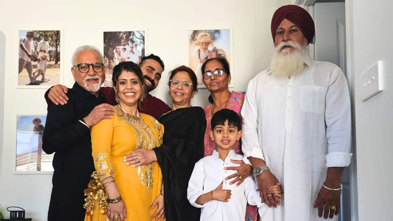A woman in a bright yellow Indian suit is surrounded by her family, some of whom are embracing her. The six adults and a young boy are posing for the camera in front of a wall with pictures in a living room.