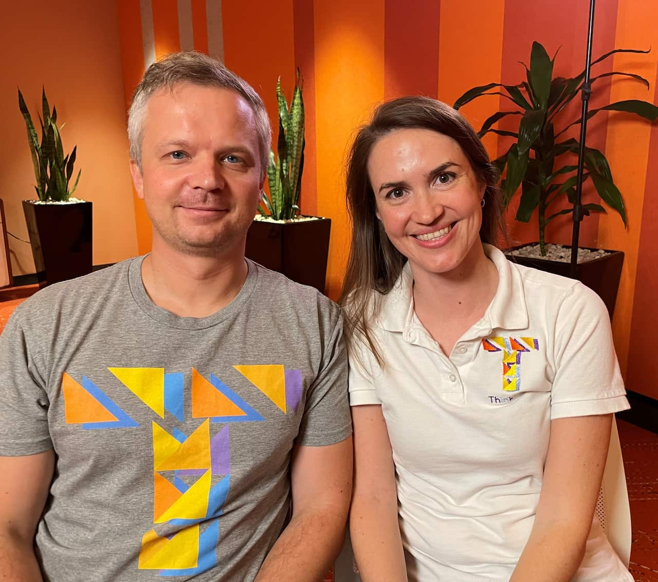 A man and a woman sitting next to each other inside and smiling. There are three pot plants behind them.